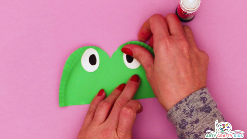 This image features hands affixing white eyes with black pupils into the green eye paper plate segment.