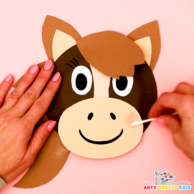 Image of a hand applying blusher to the paper plate horse to create rosy cheeks.