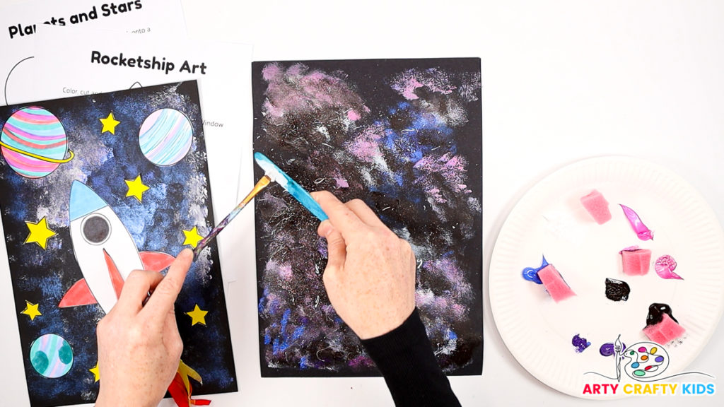 Image of a hand splatting white paint to create stars across the galaxy painting.