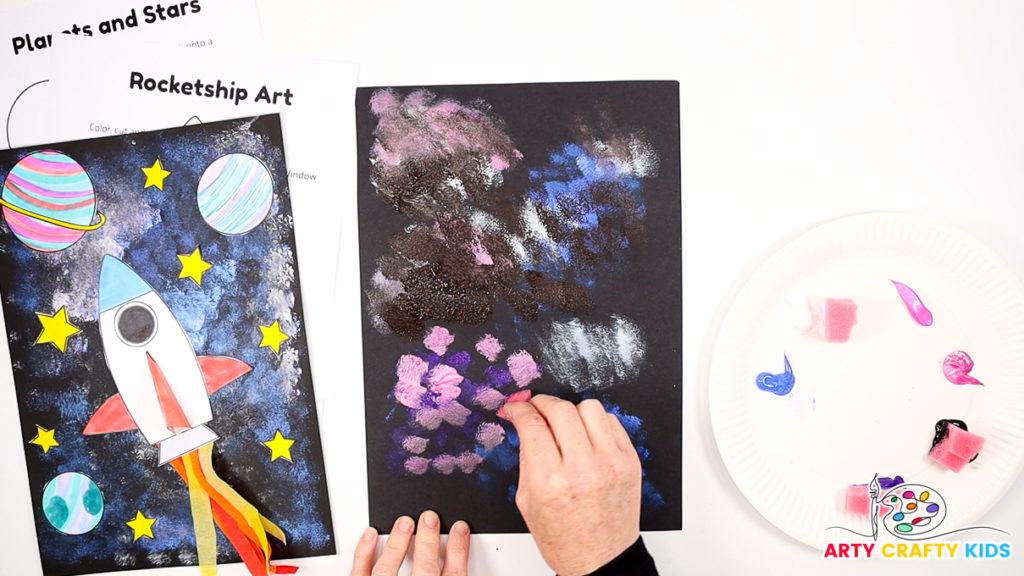 Image of a hand using a sponge to dab pink, purple and blue paint across the white to form the galaxy.