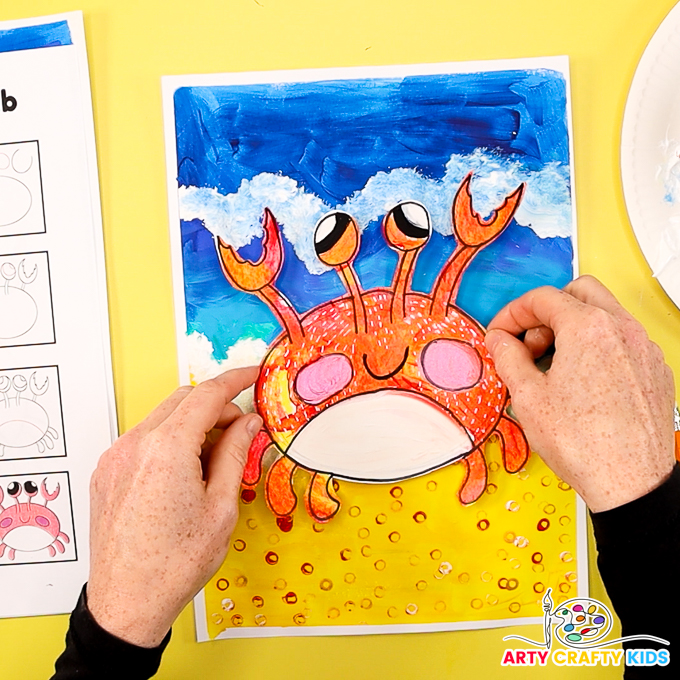 The image shows a hand affixing the crab to the sea and sand painting to complete the 'easy crab drawing and painting tutorial'.