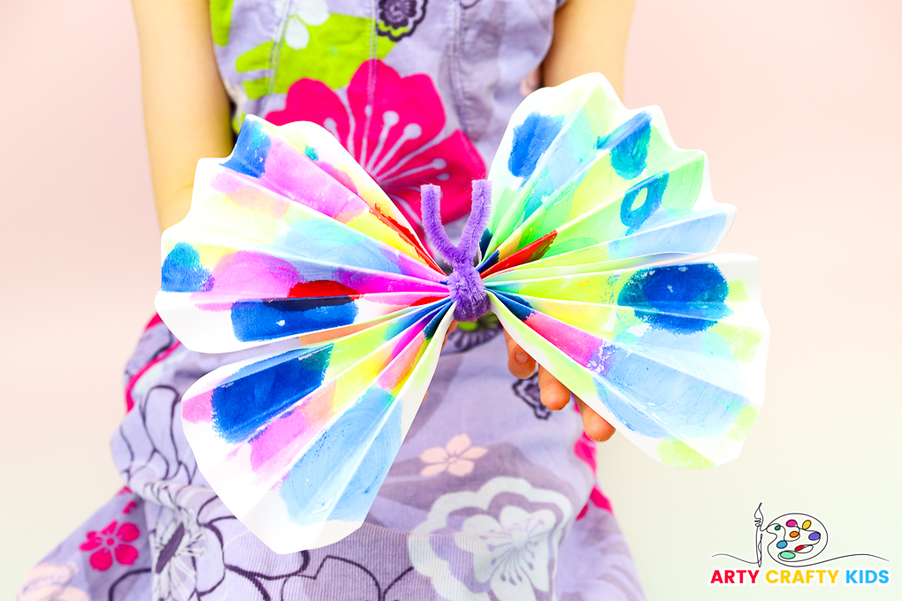 A colorful paper butterfly made from children's artwork, featuring accordion-style folded wings and a pipe cleaner body. This simple and fun craft is ideal for preschoolers, helping to recycle art and develop fine motor skills. Perfect for a vibrant school display.