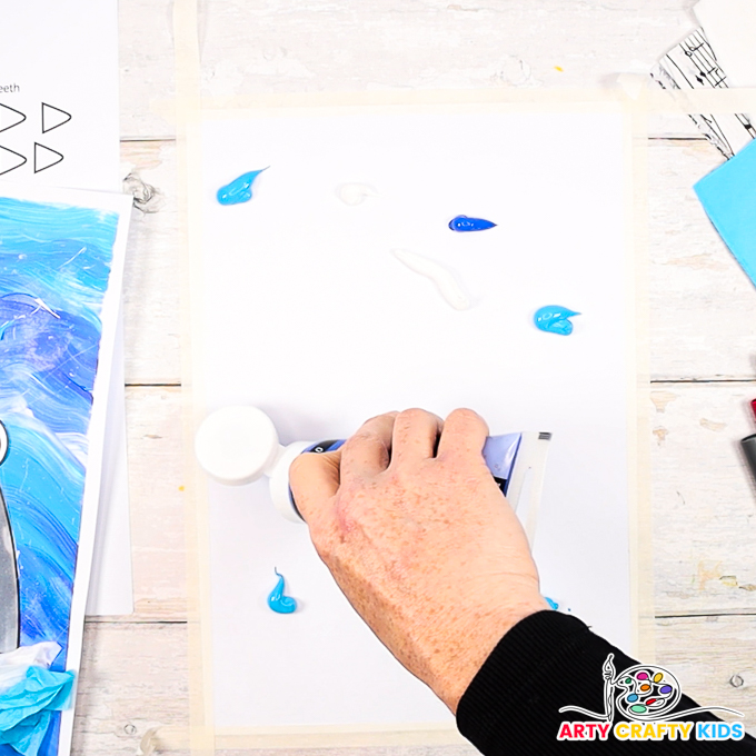 Hand squirting blue and white paint across a sheet of card stock, secured to the table top with masking tape.