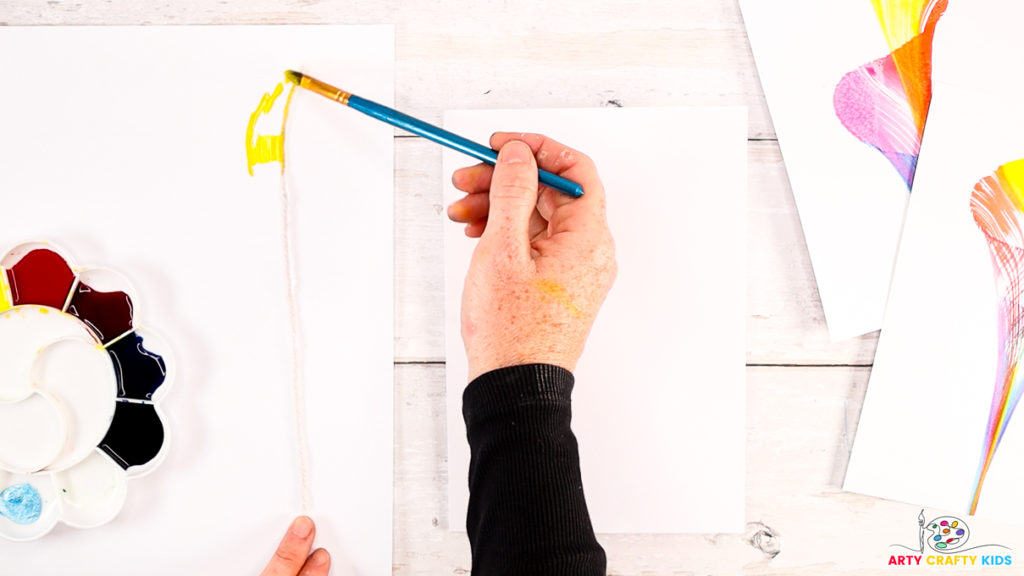 Image of step 2: A hand painting the string with watercolor paint (yellow).