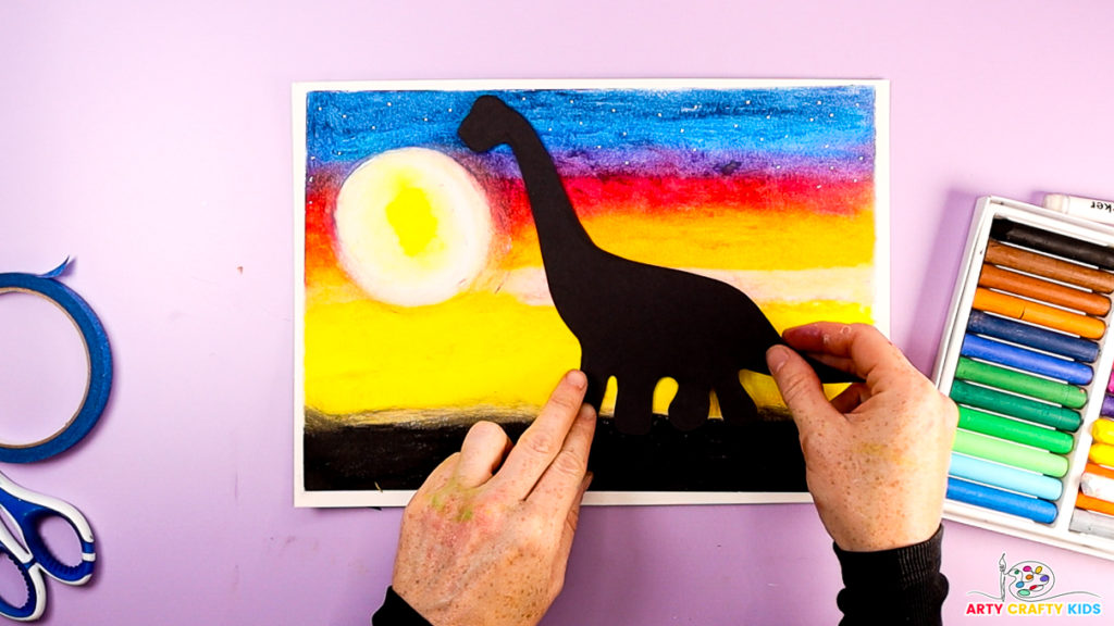 Image of a hand glueing the silhouette to the sunset to complete the sunset dinosaur silhouette art.