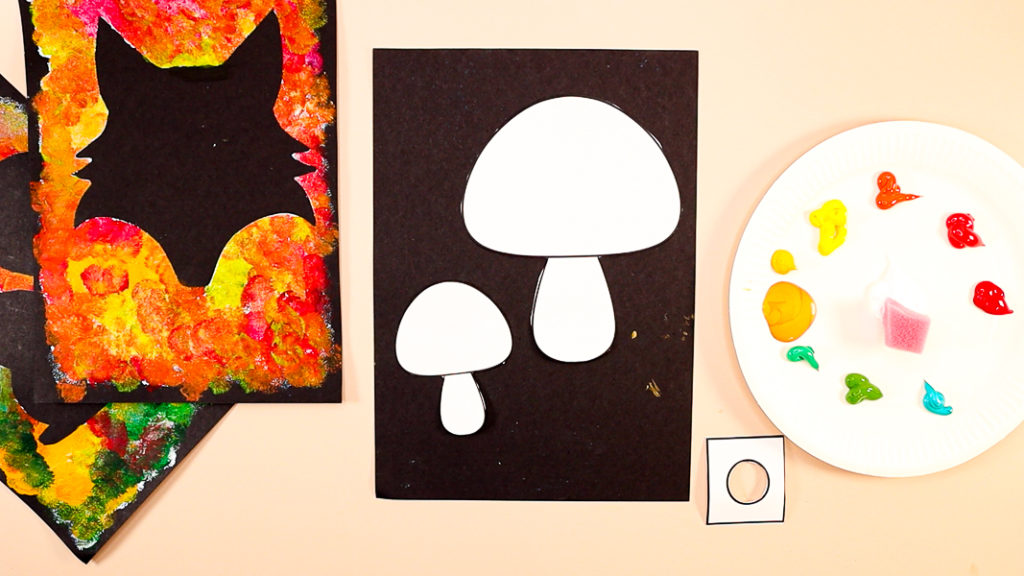 Image featuring two mushroom templates cut out and placed onto black card stock. Colorful autumn-colored paints are displayed on a paper plate with sponges for painting.