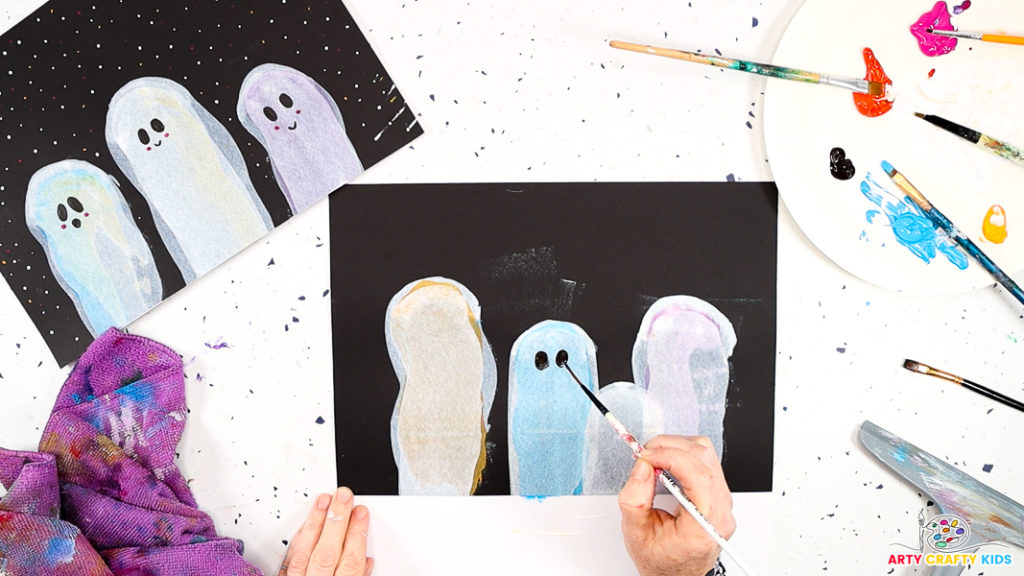 Image of a hand using a paintbrush to paint two large oval eyes onto the ghost painting.