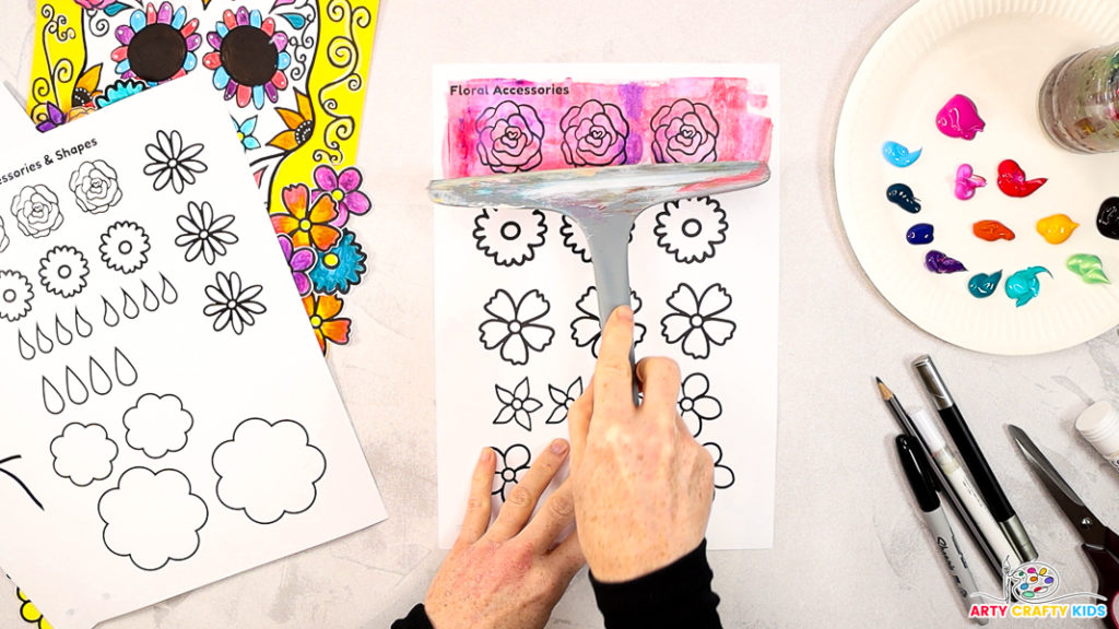 Image of a hand using a squeegee to paint the roses red, pink and purple.