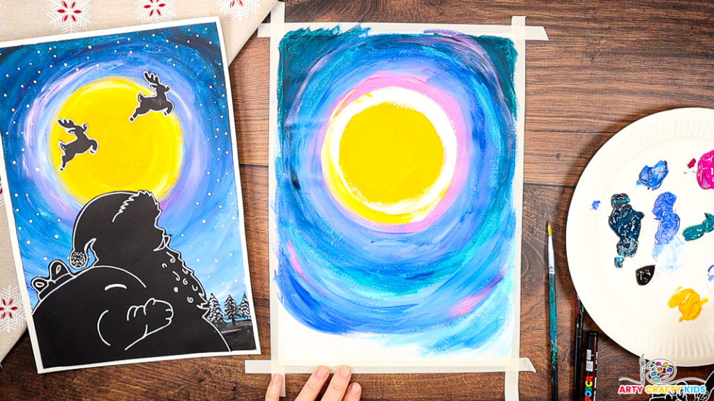 A step-by-step Christmas art project in progress, showing a vibrant painted night sky with shades of blue, pink, and purple swirling around a newly painted bright yellow moon.