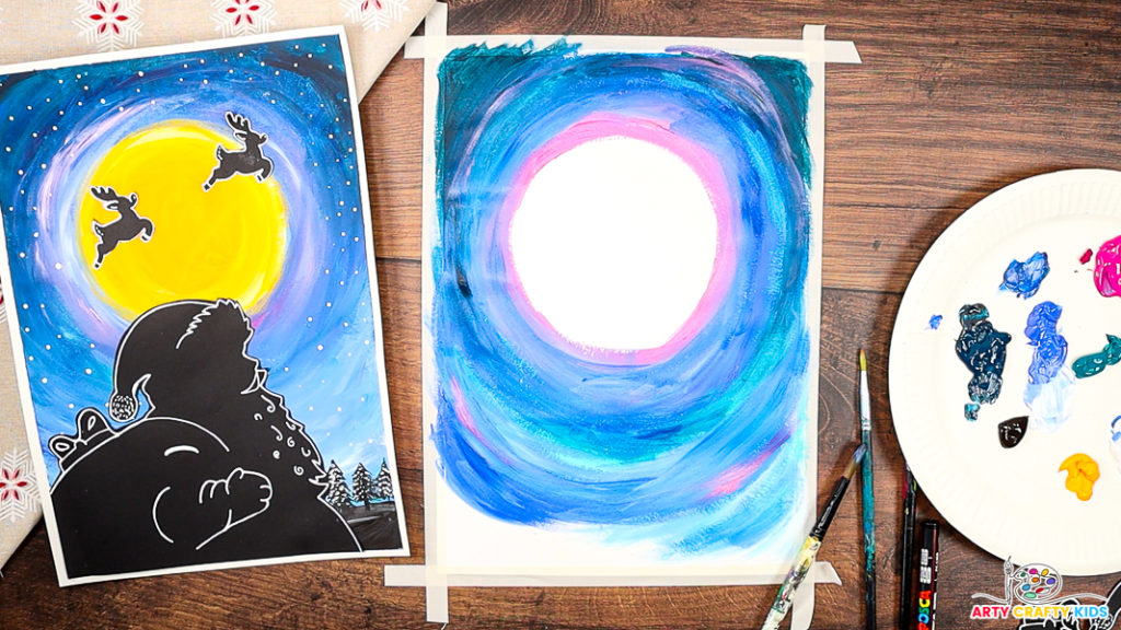 A step-by-step Christmas craft in progress, showing a partially painted night sky with layers of blue, pink, and purple swirling around a blank circular moon template. The completed Santa silhouette artwork is displayed on the left as inspiration, while a paint palette, brushes, and tools are arranged on the right, ready for use.