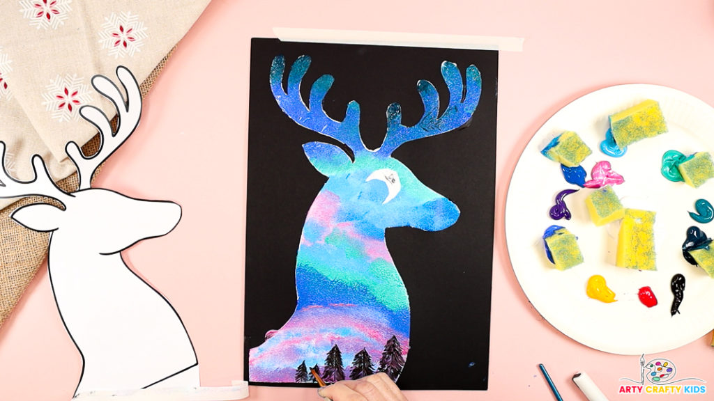 Image features a hand painting a black pine tree silhouette at the bottom of the reindeer silhouette.