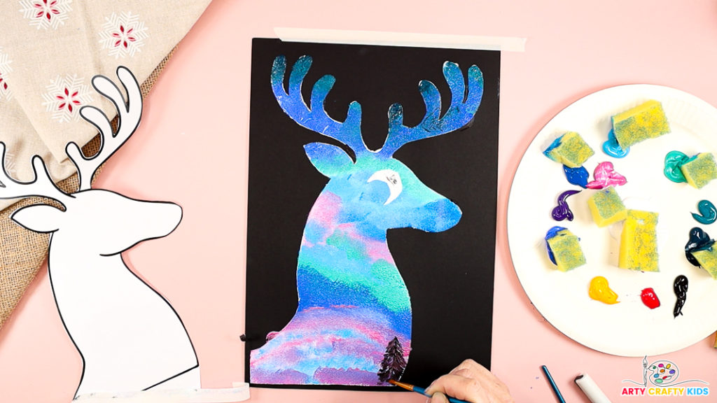 Image features a hand painting a black pine tree silhouette at the bottom of the reindeer silhouette.