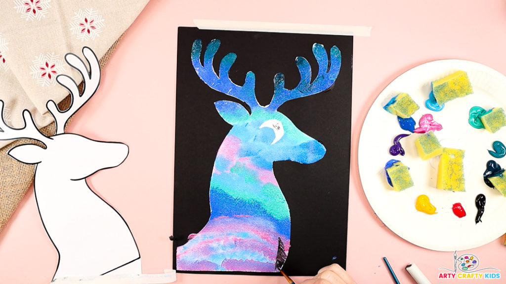 Image features a hand painting a black pine tree silhouette at the bottom of the reindeer silhouette.