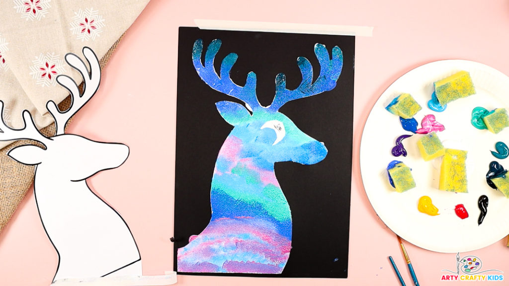 A colorful reindeer silhouette painting on black cardstock, featuring vibrant northern lights-inspired hues. A palette with paint and sponge pieces is visible alongside the completed artwork.