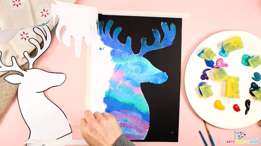 A hand carefully peels away the reindeer template, revealing a reindeer painted like the night sky.