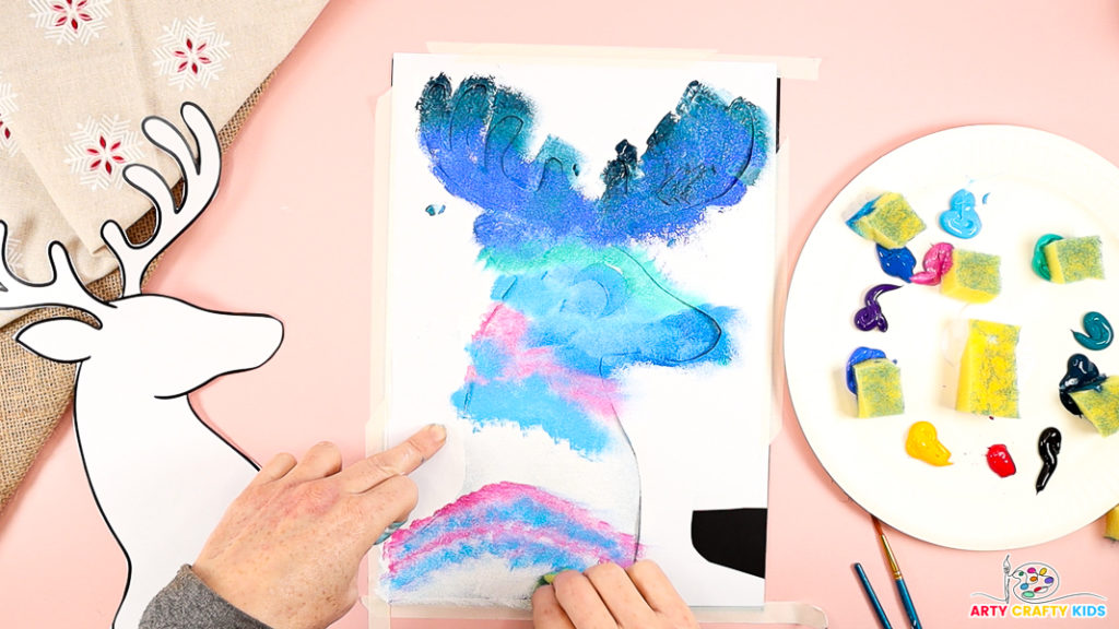 A hand is dabbing vibrant northern lights-inspired colors using a sponge onto a reindeer silhouette template. A palette with colorful paint and sponge pieces is visible on the side, along with the black reindeer cutout. The setup demonstrates a fun and creative silhouette painting process.