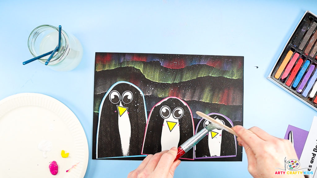 Image of a hand splattering white paint across the penguin Winter art to make a snowy effect.