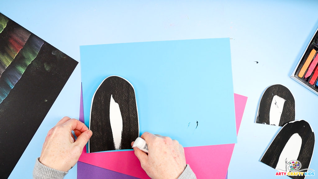 A hand is glueing a cut out penguin body to blue paper. 