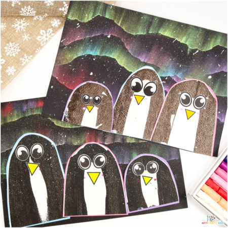 A colorful winter art project featuring scrape-painted penguins with bright yellow beaks and cartoon-style eyes. The penguins are placed on a dramatic Northern Lights background, created with blended chalk pastels and splattered white paint to resemble snowflakes.