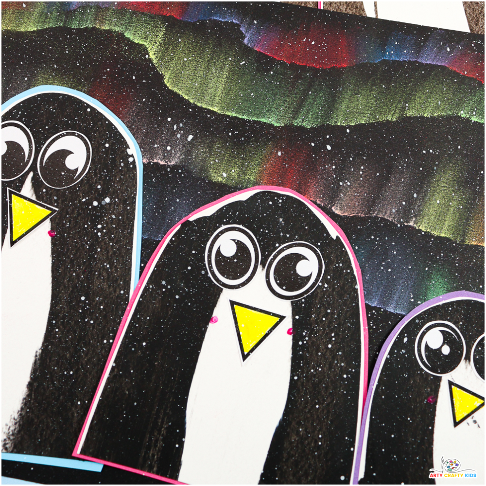 A colorful winter art project featuring scrape-painted penguins with bright yellow beaks and cartoon-style eyes. The penguins are placed on a dramatic Northern Lights background, created with blended chalk pastels and splattered white paint to resemble snowflakes.