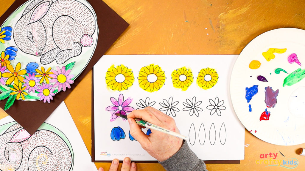 Image of a hand painting the flower template. The flowers are blue, yellow and pink.