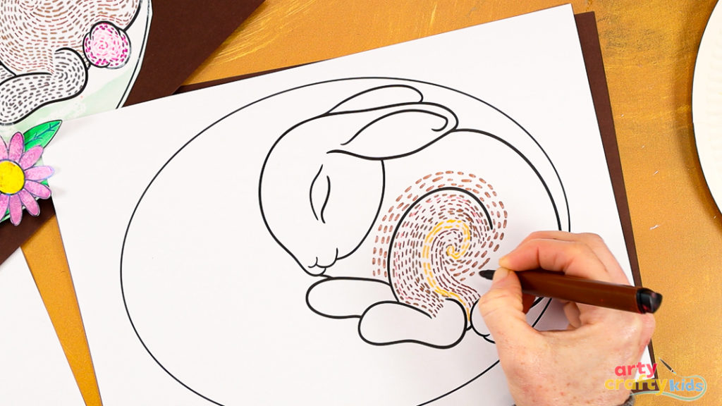 Image featuring a hand drawing a collection of dashed swirly lines within the hind leg of the sleeping bunny line art template. The lines vary in different shades of brown and yellow.