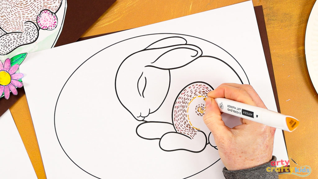 Image featuring a hand drawing a collection of dashed swirly lines within the hind leg of the sleeping bunny line art template. The lines vary in different shades of brown and yellow.