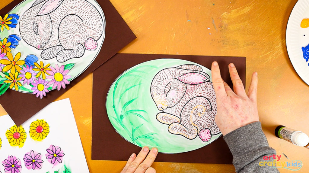 Image of a hand glueing a cut out sleeping bunny line art onto brown paper.