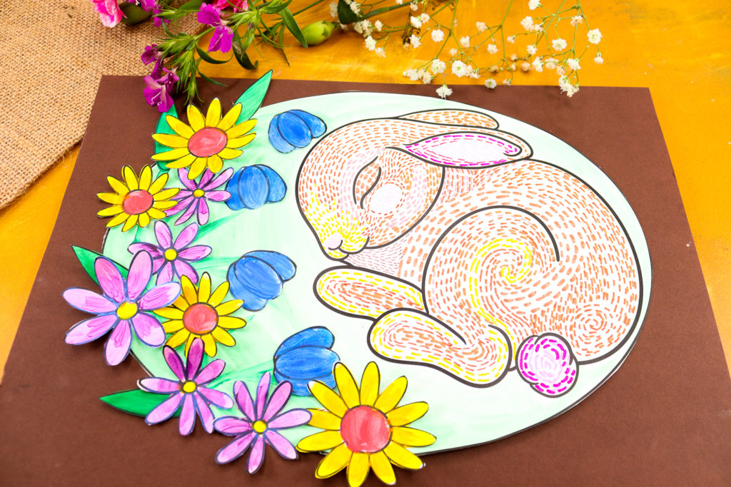 completed sleeping bunny line art project, cut into an oval shape and glued onto reddish-brown cardstock, creating the effect of a bunny nestled inside its burrow. Vibrant flowers add a pop of color and depth.