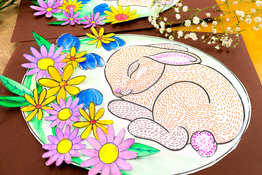 completed sleeping bunny line art project, cut into an oval shape and glued onto reddish-brown cardstock, creating the effect of a bunny nestled inside its burrow. Vibrant flowers add a pop of color and depth.