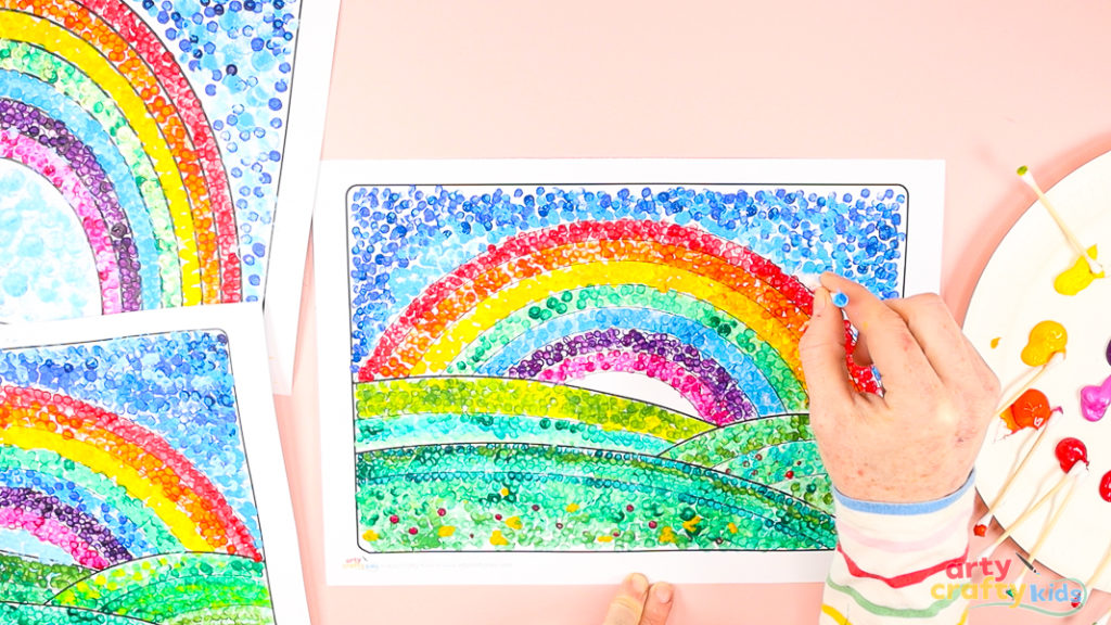 An in-progress pointillism rainbow painting, with a hand dotting green paint onto the landscape. A plate of mixed paint colors and Q-tips sit nearby, ready to add more detail.