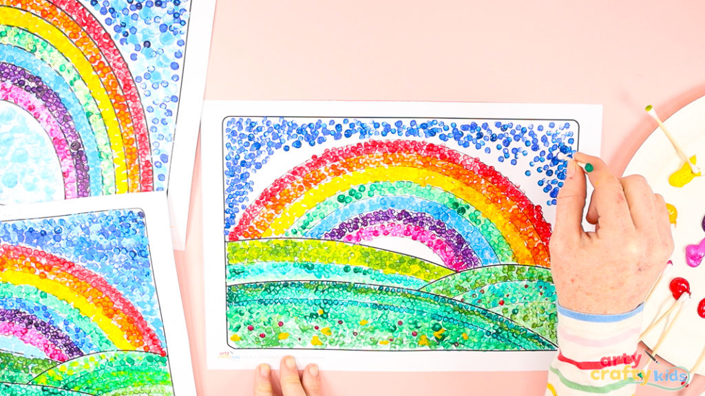 Image featuring a hand filling the space around the completed rainbow with blue dots.